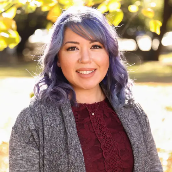 A woman with wavy, shoulder-length purple hair smiles outdoors. She wears a maroon lace top and a gray cardigan, with sunlight and blurred yellow leaves in the background.