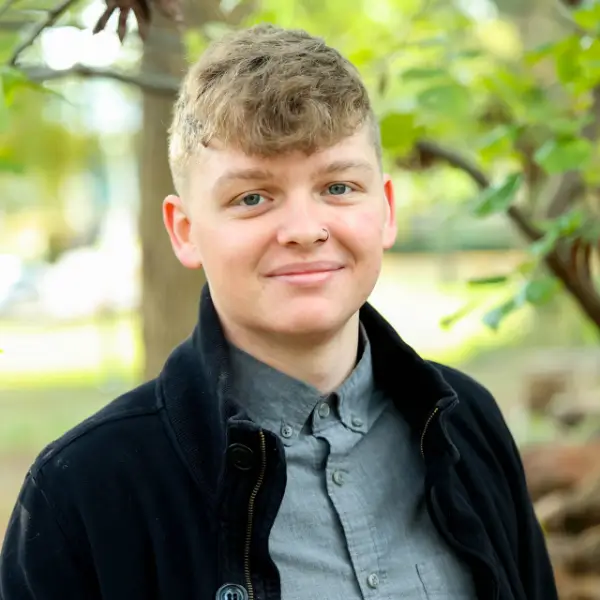 A young person with short, curly light brown hair, wearing a gray button-up shirt and a dark jacket, stands outside in a park-like setting with green trees in the background, smiling at the camera.