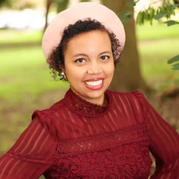 A woman with curly hair wearing a light pink beret and a burgundy lace top smiles at the camera while standing outdoors with greenery in the background.