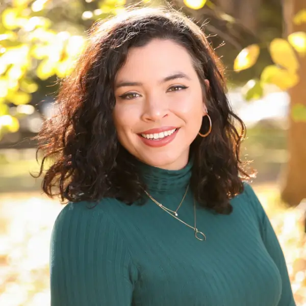 A woman with wavy dark hair, wearing a green ribbed turtleneck and gold hoop earrings, smiles outdoors in a sunlit park with blurred autumn foliage in the background.