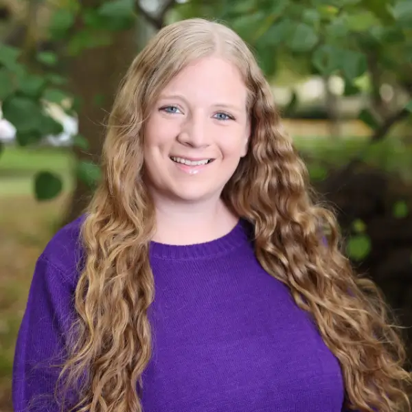 A woman with long, wavy blonde hair wearing a purple sweater smiles outdoors, with green foliage in the background.