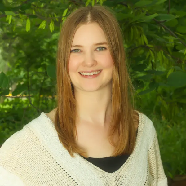A young woman with straight, light brown hair smiles at the camera. She is wearing a cream-colored sweater and standing outdoors in front of leafy green trees.