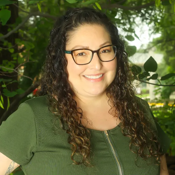 A woman with long curly hair and glasses smiles outdoors, wearing a short-sleeved green top with a zippered front. Lush green foliage fills the background.