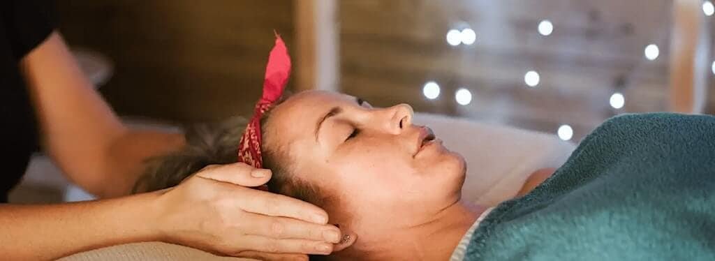 A person with a red headband lies on a massage table, eyes closed, while another person gently massages their head. Soft lighting and string lights create a relaxing atmosphere.
