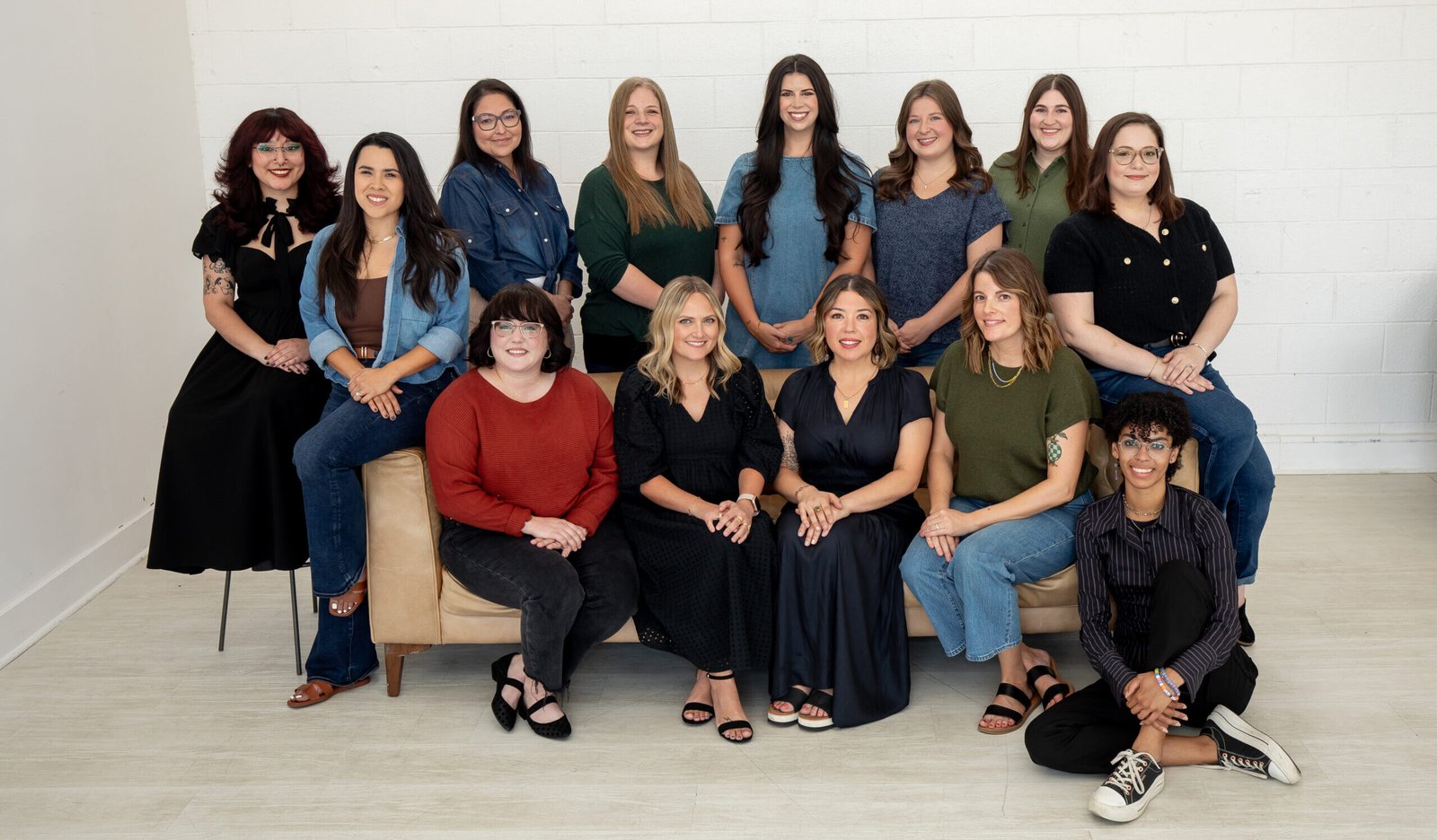 A group of fourteen women pose together indoors. Some are sitting on a tan couch or on the floor, while others stand behind it. They are smiling and casually dressed. The background is a white wall and light wood floor.