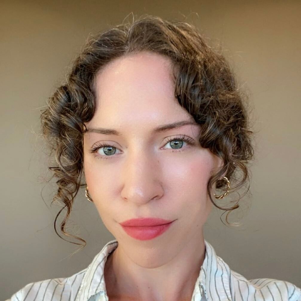 A person with curly brown hair, light eyes, and wearing gold hoop earrings and a white shirt with thin stripes, poses in front of a neutral background.